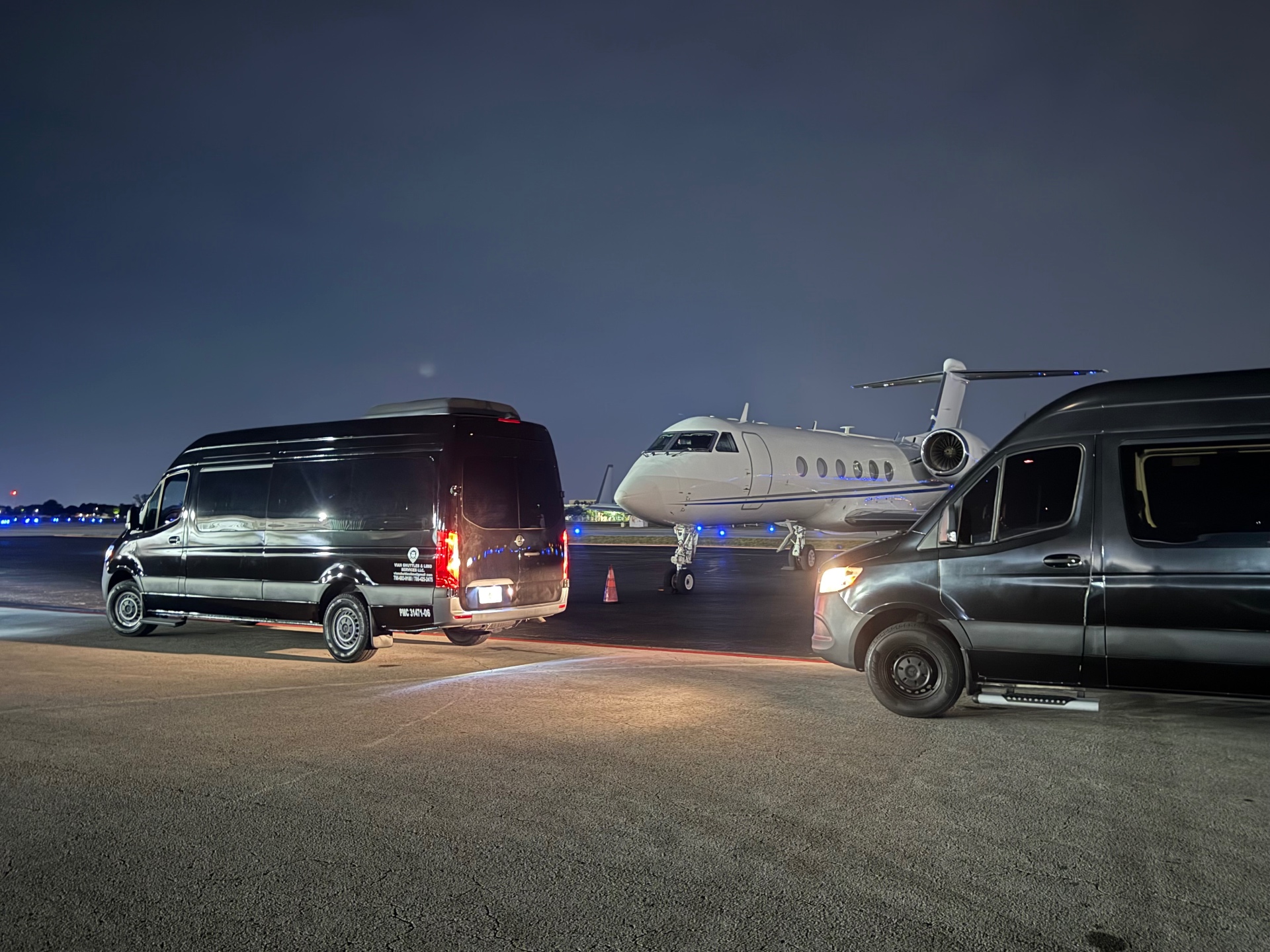 Two black Vian Sprinter vans on the tarmac beside a private jet at dusk